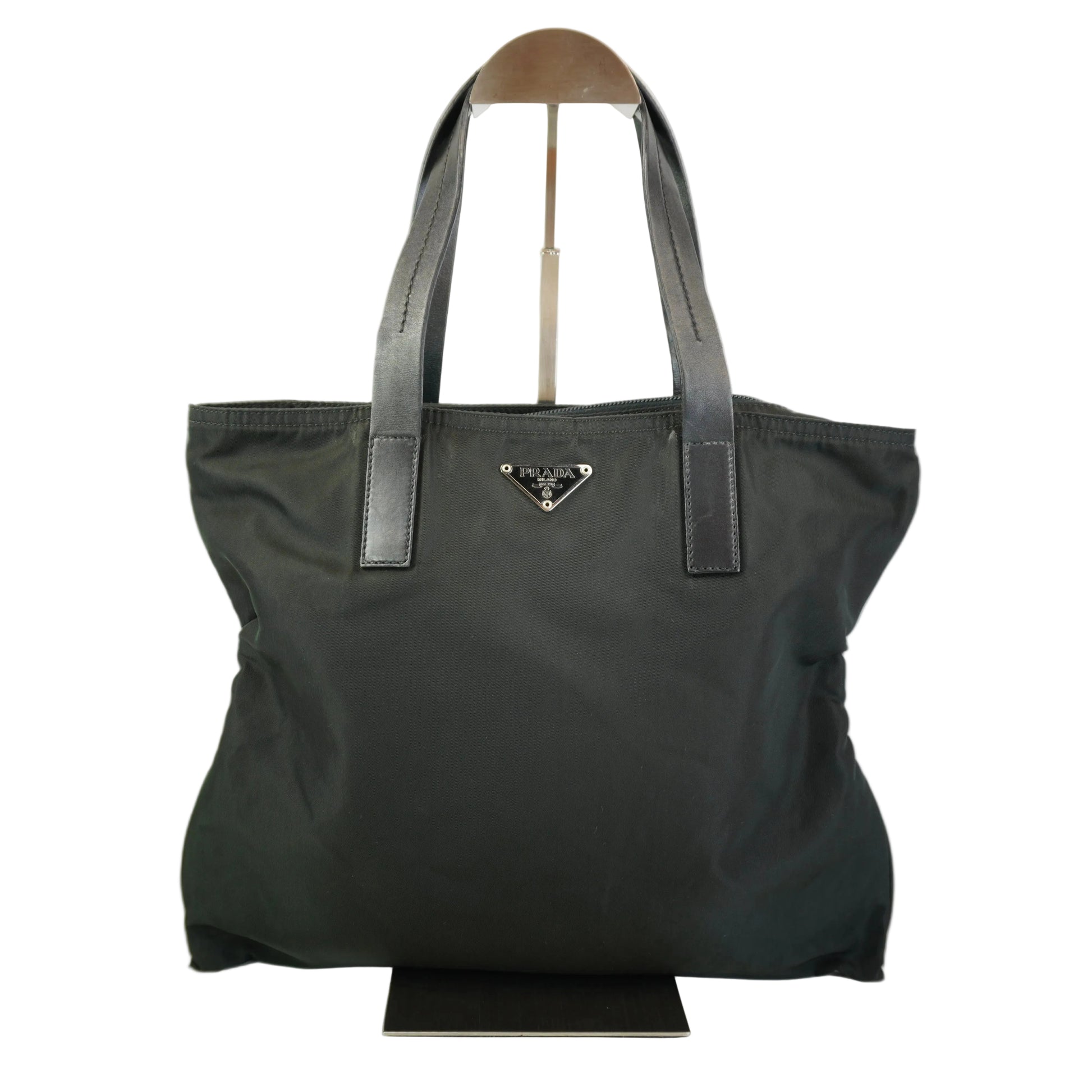 A black Prada nylon tote bag with dark gray leather shoulder straps and a silver-tone triangular logo plaque on the front, displayed on a stand against a white background.