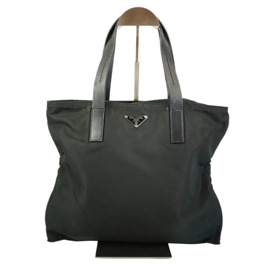A black Prada nylon tote bag with dark gray leather shoulder straps and a silver-tone triangular logo plaque on the front, displayed on a stand against a white background.